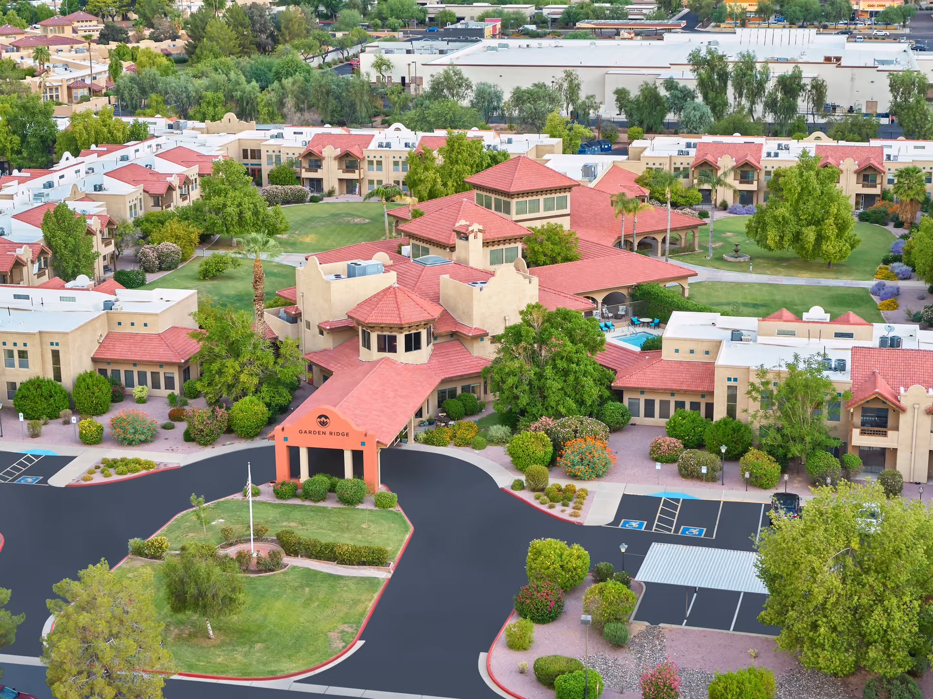Aerial view of Garden Ridge senior living facility featuring multiple beige buildings with red-tiled roofs, surrounded by green lawns, trees, and landscaped bushes. The main entrance has a covered driveway with the Garden Ridge logo and name displayed. Parking spaces, including handicapped spots, are visible near the entrance.