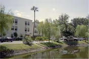 Light-colored senior living building beside a pond with trees and parked cars.