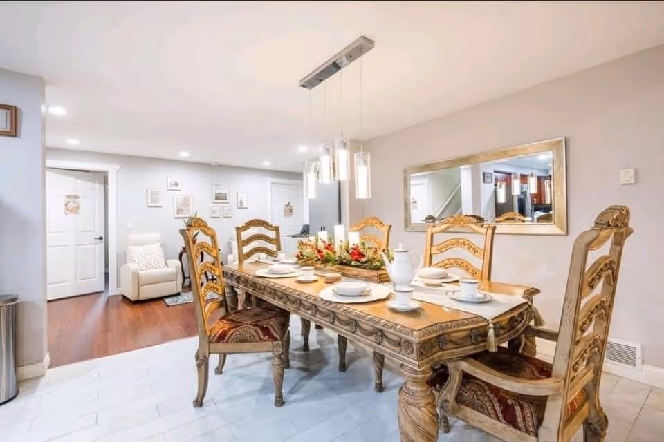 A well-lit dining room with a wooden dining table set for six people. The table has ornate carved legs and matching chairs with patterned cushions. A centerpiece with candles and flowers decorates the table. A large rectangular mirror hangs on the wall reflecting part of the adjacent kitchen. The floor is tiled near the dining area and transitions to wood flooring in the background where a white armchair is visible. The walls are painted light gray and recessed ceiling lights illuminate the space.