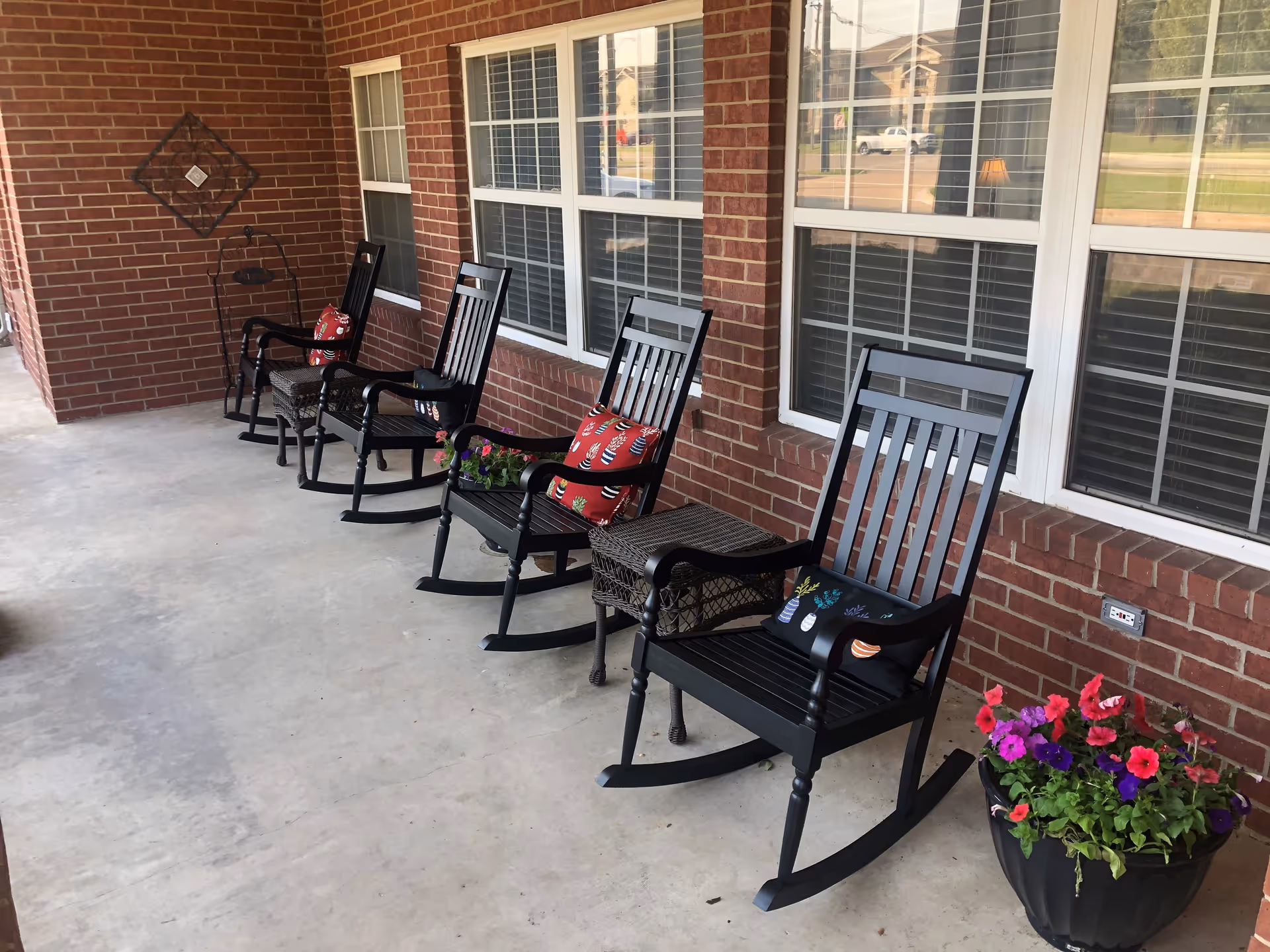 A covered outdoor porch area with four black rocking chairs, each with a decorative cushion. There are two small wicker tables between the chairs and a large flower pot with colorful flowers on the right side. The porch has a brick wall with several windows and a concrete floor.