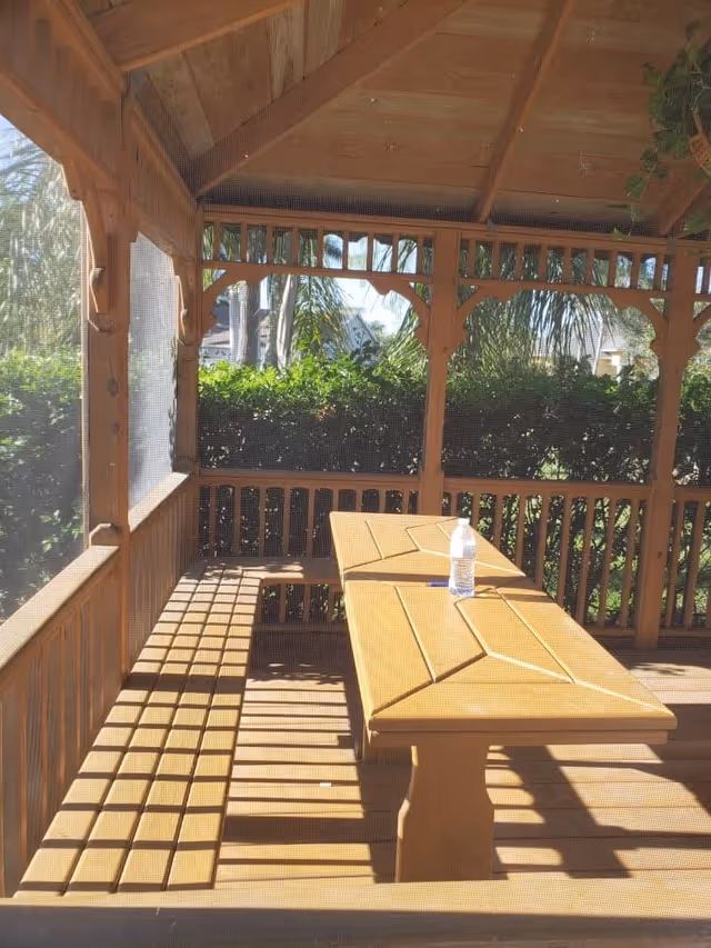 Wooden gazebo with a picnic table and benches inside, surrounded by greenery and palm trees visible through the open sides.
