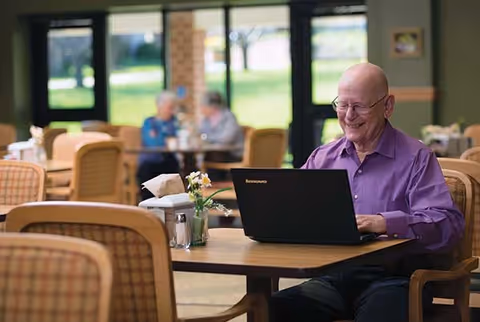 An elderly man wearing a purple shirt is sitting at a table in a dining area, smiling while using a Lenovo laptop. The table has a small vase with flowers and some condiments. In the background, two elderly women are seated and engaged in conversation near large windows showing a green outdoor area.
