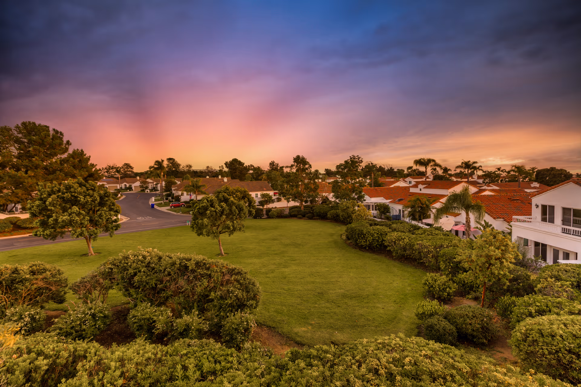 A scenic view of a residential community at sunset with a well-maintained green lawn, various trees and bushes, and houses with red-tiled roofs under a colorful sky transitioning from orange to purple.