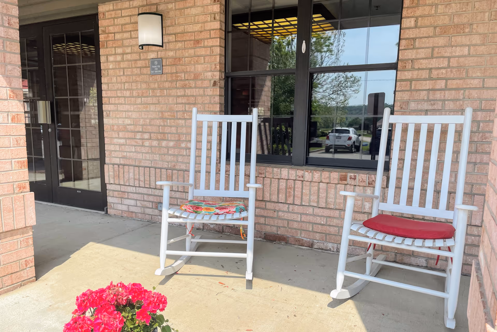 Two white wooden rocking chairs with cushions on a concrete porch outside a brick building. A window and a glass door are visible behind the chairs, along with a wall-mounted light fixture and a small sign. A pot of bright pink flowers is in the foreground on the porch.