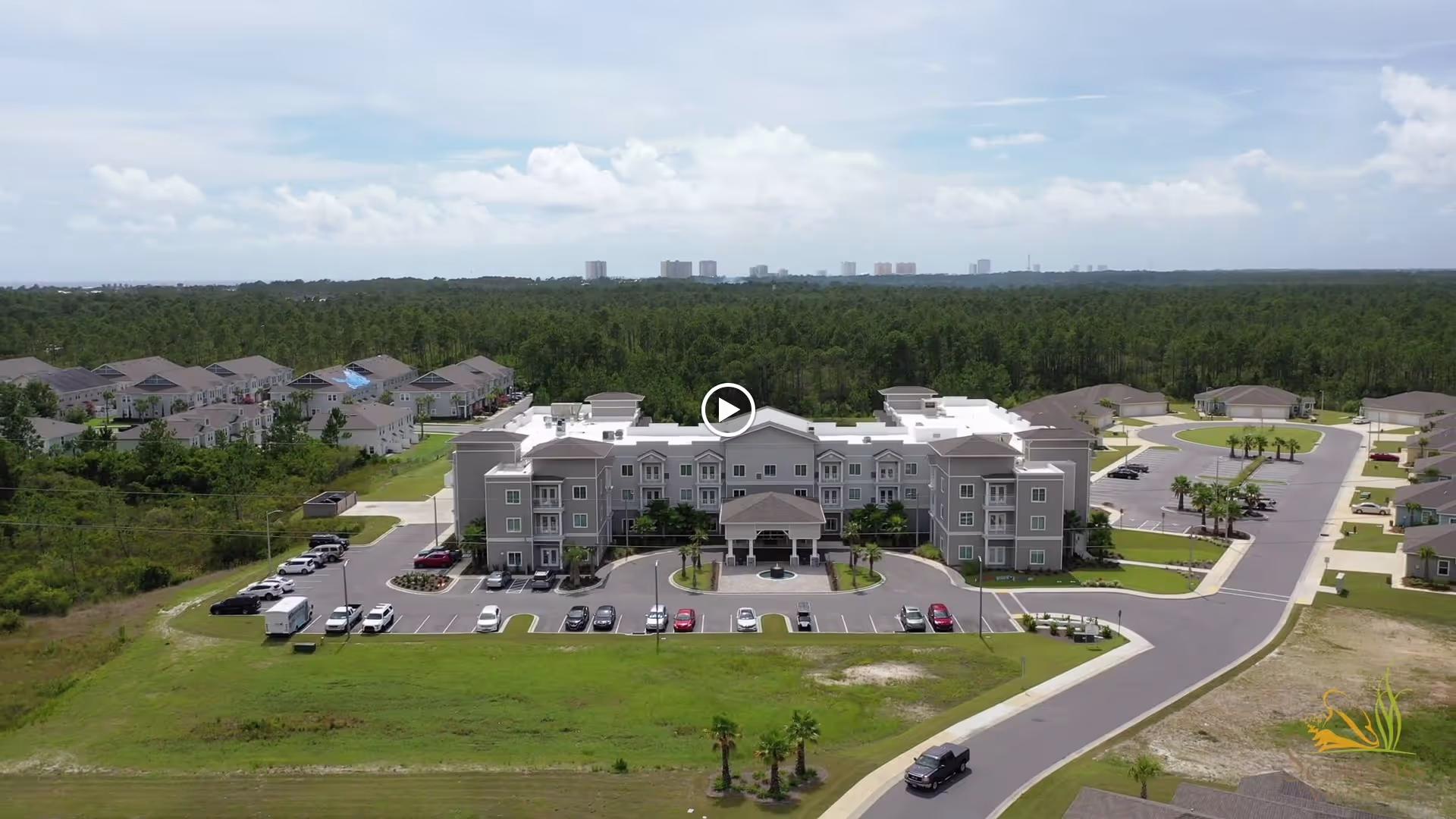 Aerial view of a three-story senior living facility with a circular driveway, parked cars, surrounding lawns and nearby homes.