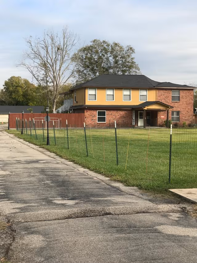 Exterior view of a two-story brick and yellow siding building with a black roof, surrounded by a green lawn and a wire fence. There is a paved driveway leading up to the building, and trees are visible in the background under a cloudy sky.