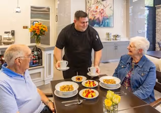A smiling chef serves coffee to two elderly residents seated at a dining table with bowls of cereal and fruit in a bright dining area.