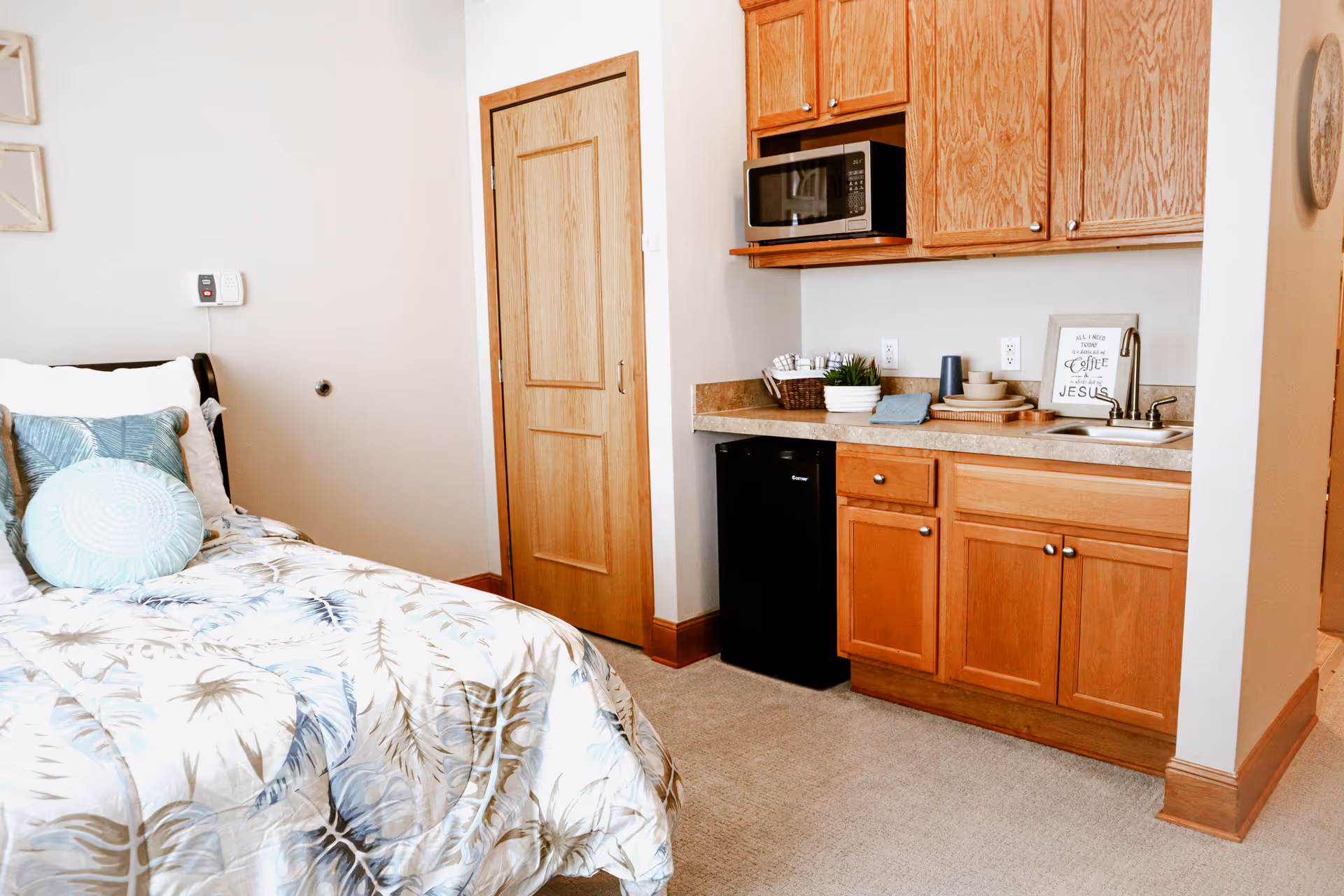 A cozy room featuring a bed with patterned bedding and decorative pillows on the left, and a kitchenette area on the right with wooden cabinets, a microwave, a small refrigerator, a sink, and countertop accessories including a basket, bowls, and a framed sign.
