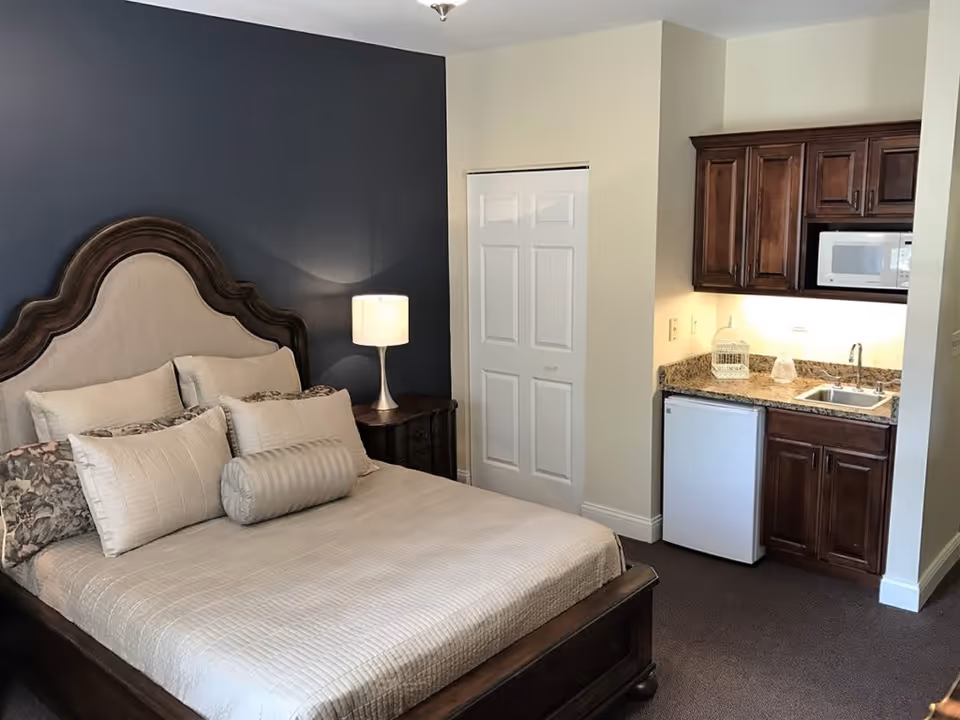 A cozy bedroom with a large bed featuring a decorative wooden headboard and multiple pillows. Next to the bed is a nightstand with a lit table lamp. In the corner of the room, there is a small kitchenette area with dark wooden cabinets, a granite countertop, a mini refrigerator, a microwave, and a sink. The walls are painted in a combination of dark blue and light beige.