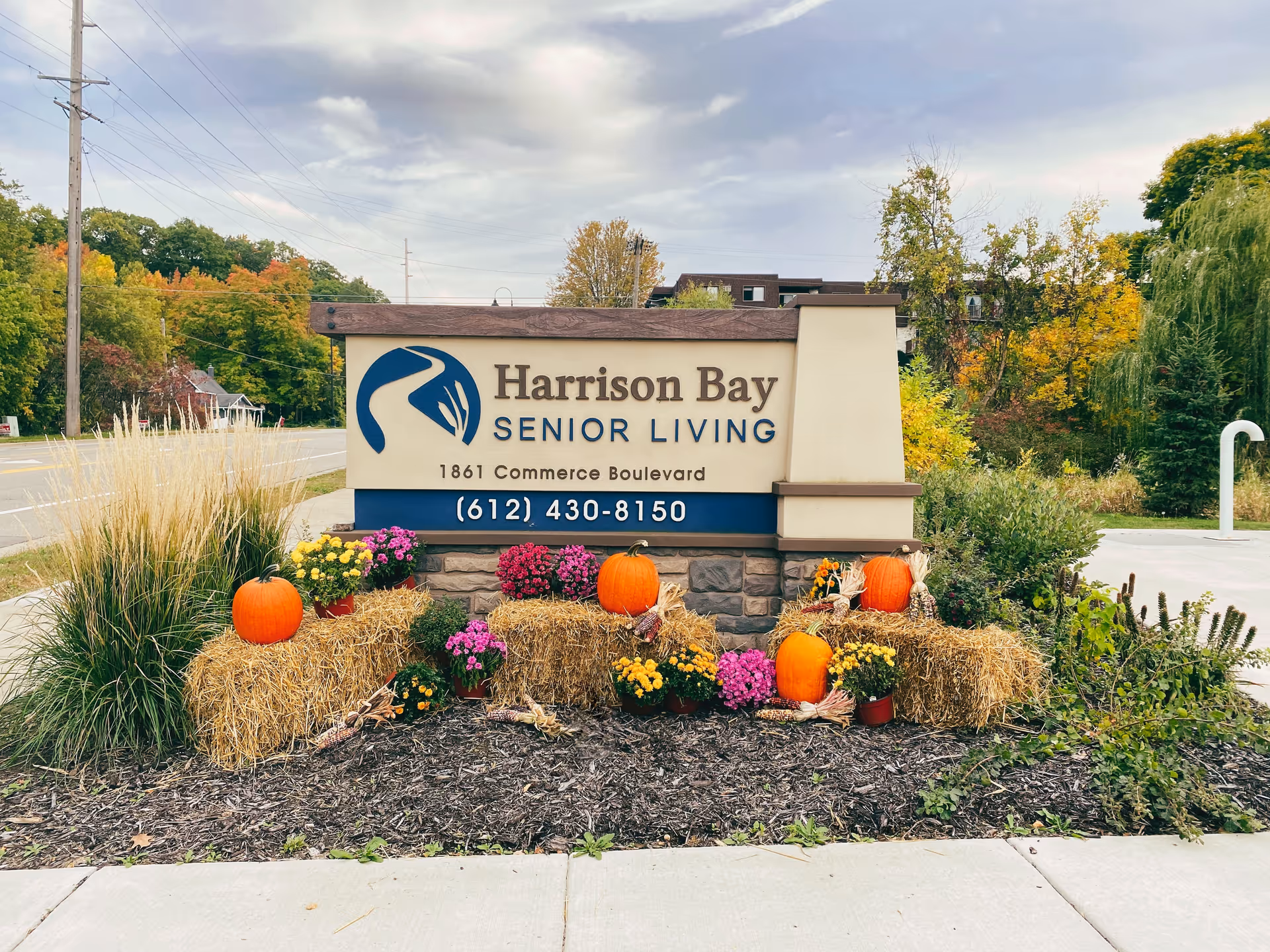 Outdoor sign for Harrison Bay Senior Living at 1861 Commerce Boulevard, decorated with pumpkins, hay bales, and colorful flowers, with trees and a road in the background under a cloudy sky.