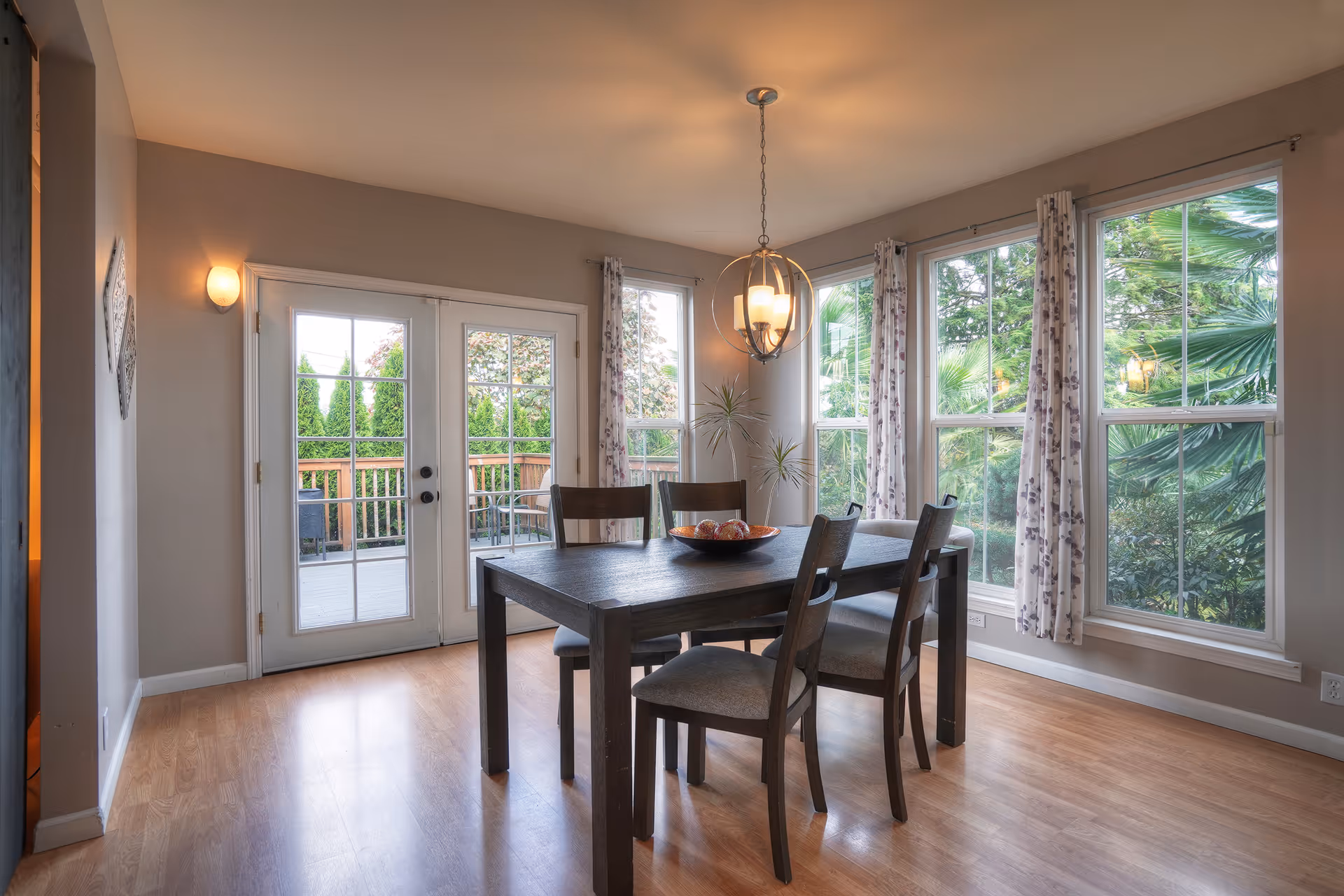 Bright dining room with a dark wood table and four chairs, large windows and French doors opening to a deck.