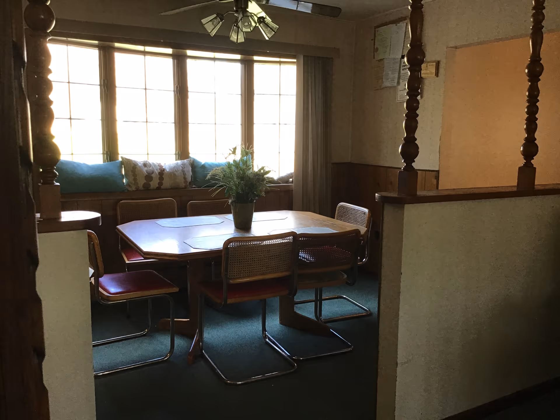 A dining area with a wooden table surrounded by six chairs with red cushions and cane backs. A potted plant is placed in the center of the table. Behind the table is a large window with multiple panes, a window seat with blue and patterned pillows, and curtains on the side. The room has wood paneling on the lower half of the walls and a ceiling light fixture with multiple bulbs.
