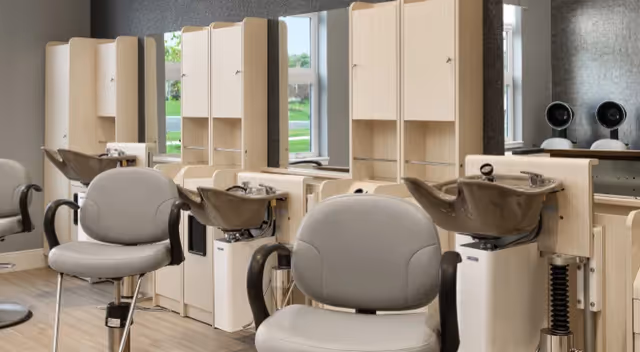 Salon area with gray styling chairs, shampoo bowls, mirrors, and hooded hair dryers.