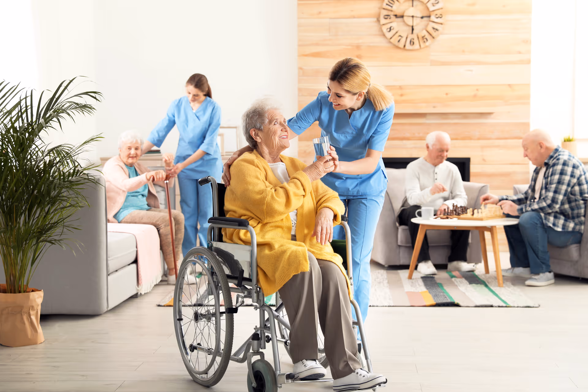 A caregiver in blue scrubs is helping an elderly woman in a yellow cardigan sitting in a wheelchair by giving her a glass of water. In the background, another caregiver assists an elderly woman sitting on a couch, while two elderly men play chess at a small table in a cozy living room with wooden paneling and a large clock on the wall.