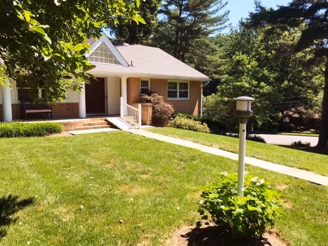 A single-story brick building with a front porch supported by white columns, surrounded by green grass and trees. A concrete walkway leads to the porch, and there is a small garden lamp post in the foreground.