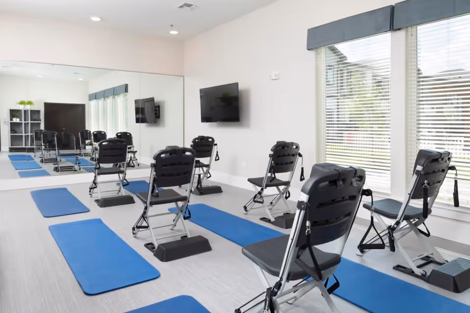 A bright exercise room with several black folding chairs equipped with resistance bands, each placed on blue exercise mats. The room has a large wall mirror, a mounted flat-screen TV, and windows with blinds allowing natural light to enter.