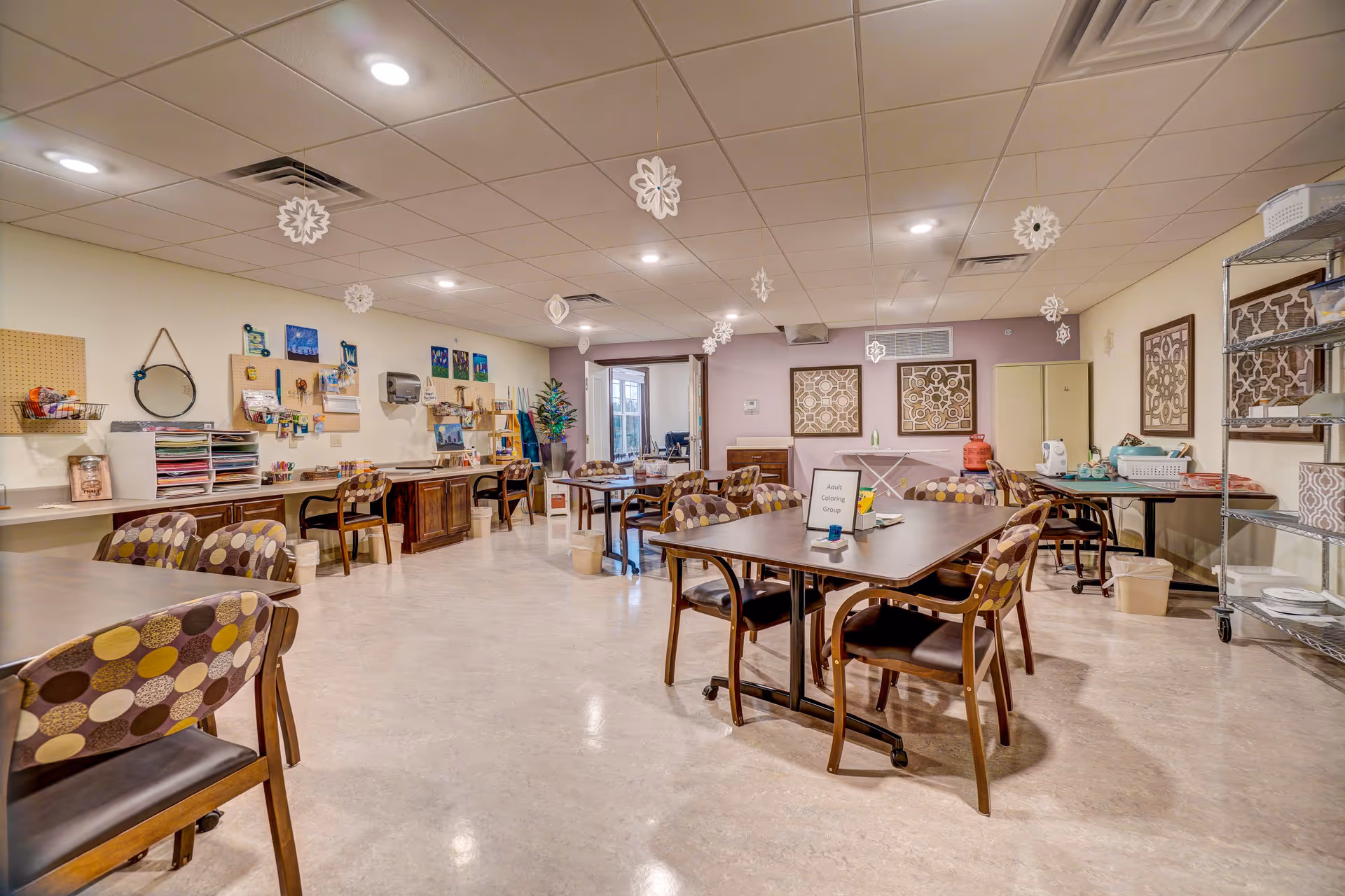 A spacious activity room with several tables and chairs arranged for group activities. The chairs have patterned upholstery with circular designs. The walls are decorated with artwork and pegboards holding craft supplies. Snowflake decorations hang from the ceiling. A sign on one table reads 'Adult Coloring Group'. Shelving units and various craft materials are visible around the room.