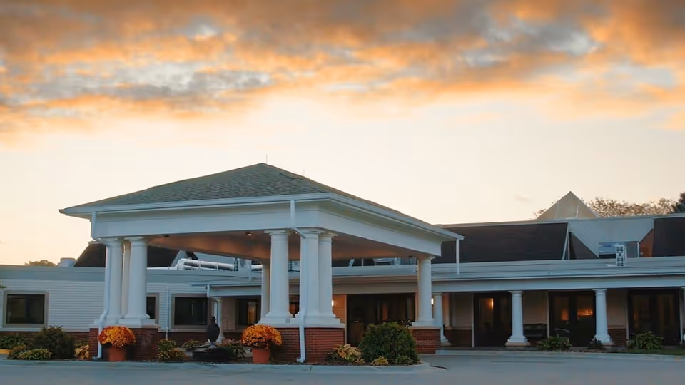 Exterior view of a senior living facility building with a covered entrance supported by white columns, potted plants with orange flowers near the entrance, and a sunset sky in the background.