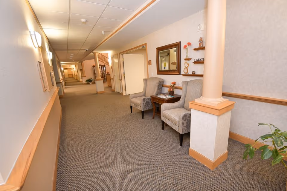 Carpeted interior hallway in a senior living facility with a small seating area of two chairs, a side table and decorative shelves.