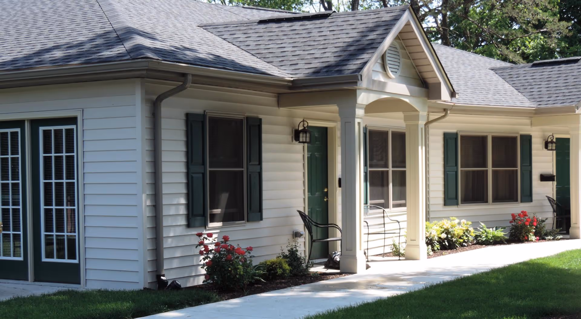 Front exterior of a single-story senior living building with a small covered entrance, green doors and shutters, and a landscaped walkway.