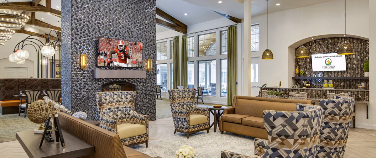 Spacious senior living community lounge with patterned armchairs and sofas arranged around a central stone fireplace wall and a welcome screen above a bar area.