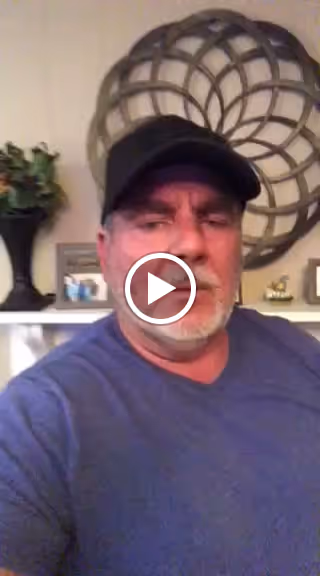 A man wearing a black cap and a blue shirt is sitting indoors. Behind him, there is a decorative circular wall piece, a small plant on a stand, and some framed photos on a shelf.