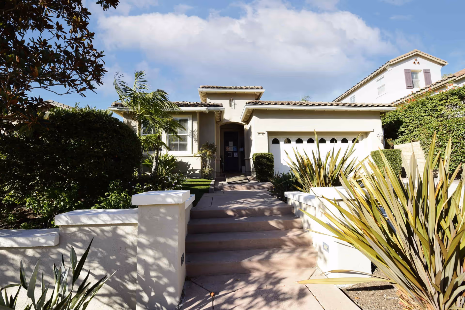 Front exterior view of a single-story residential building with a tiled roof, a garage, and a walkway leading up to the entrance. The house is surrounded by various plants and shrubs under a partly cloudy sky.