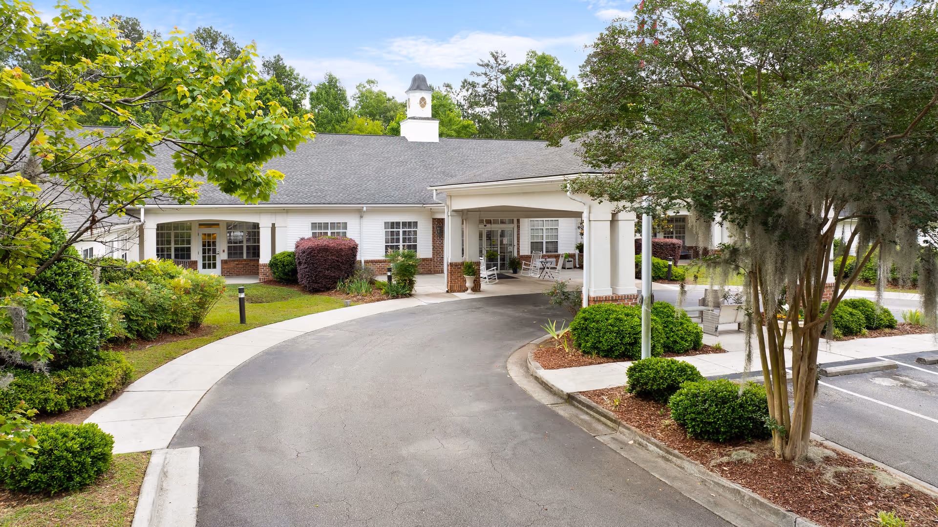 Exterior view of TerraBella Summerville facility showing a curved driveway leading to a covered entrance with white columns. The building has white siding with brick accents and a clock tower on the roof. Surrounding the driveway are well-maintained shrubs, trees, and landscaped areas with a parking lot visible on the right side.