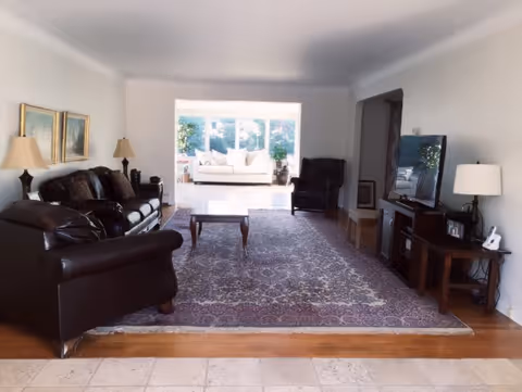 A spacious living room with dark leather sofas and armchairs arranged around a wooden coffee table on a large patterned rug. The room has light-colored walls with two framed paintings and two table lamps on side tables. In the background, a bright sunroom with a white sofa and large windows letting in natural light is visible.
