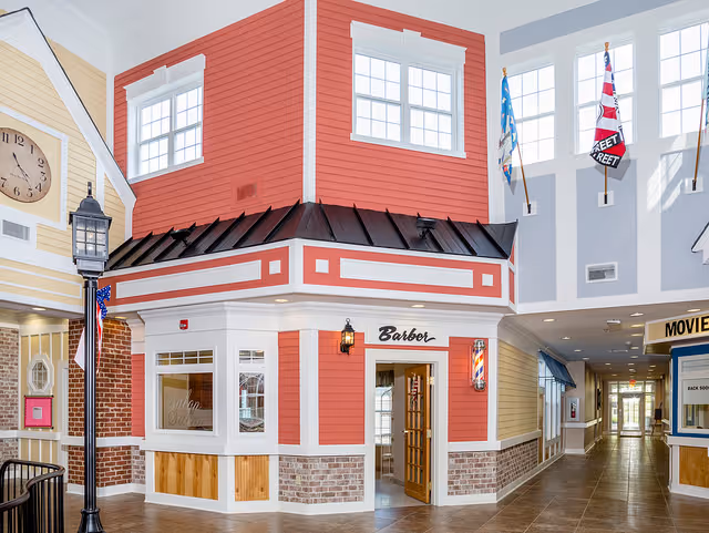 Interior view of a senior living facility designed to look like a small town street with a barber shop featuring red and white exterior walls, a black roof, and a traditional barber pole. The area has large windows, a street lamp, and flags mounted on the walls. A hallway extends to the right with additional storefront-style facades and a movie theater entrance visible.