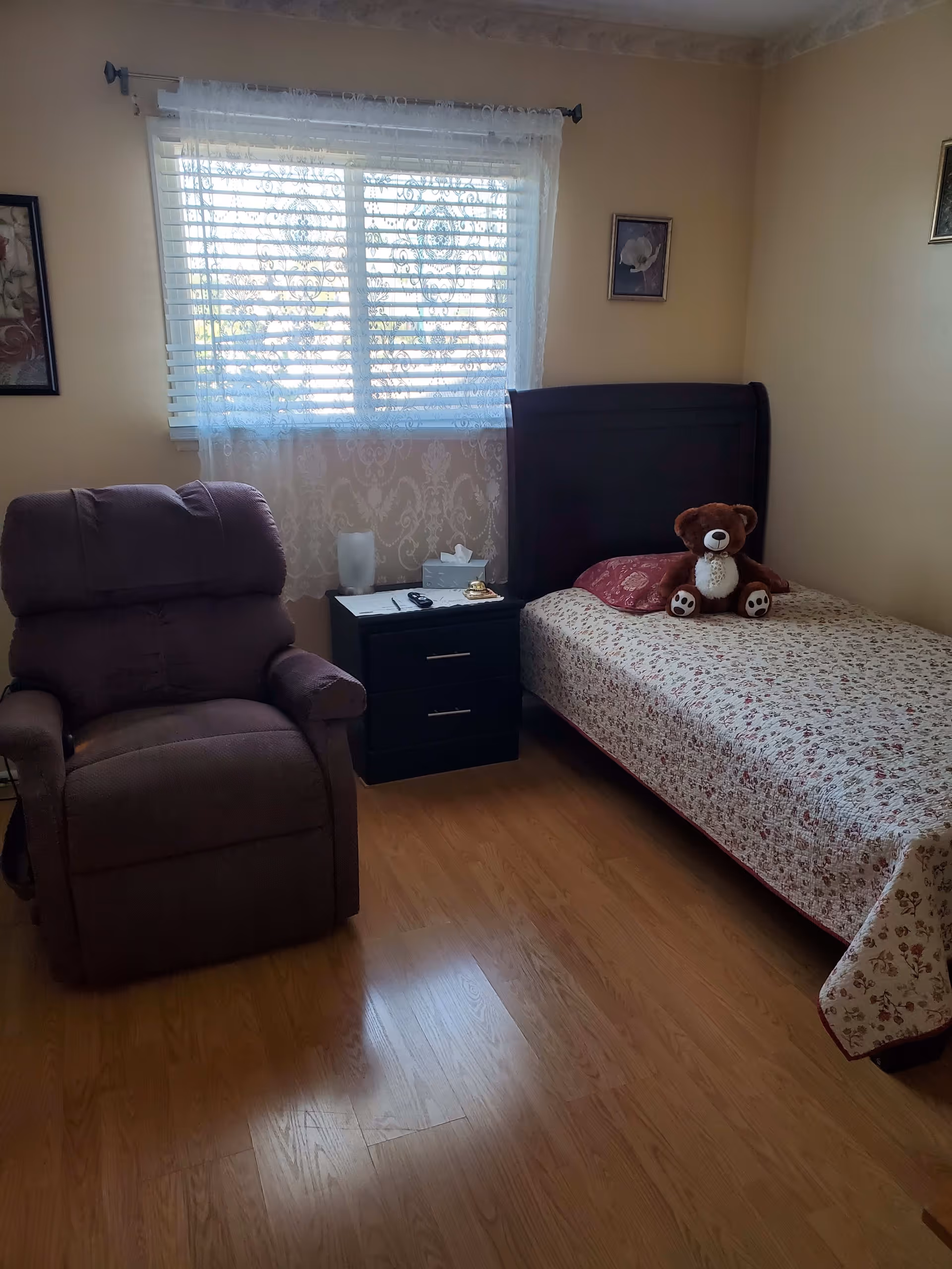 A cozy bedroom with a single bed covered in a floral quilt, a brown teddy bear sitting on the bed, a dark wooden headboard, a nightstand with a lamp, tissue box, remote control, and a small bell. Next to the nightstand is a comfortable brown recliner chair. The room has light-colored walls, a window with sheer white curtains and blinds, and two framed pictures hanging on the walls.