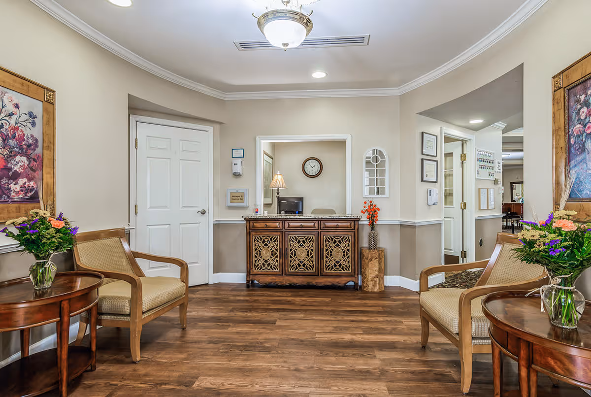 Reception area with two chairs, side tables holding flower arrangements, and a decorative reception desk under a ceiling light.