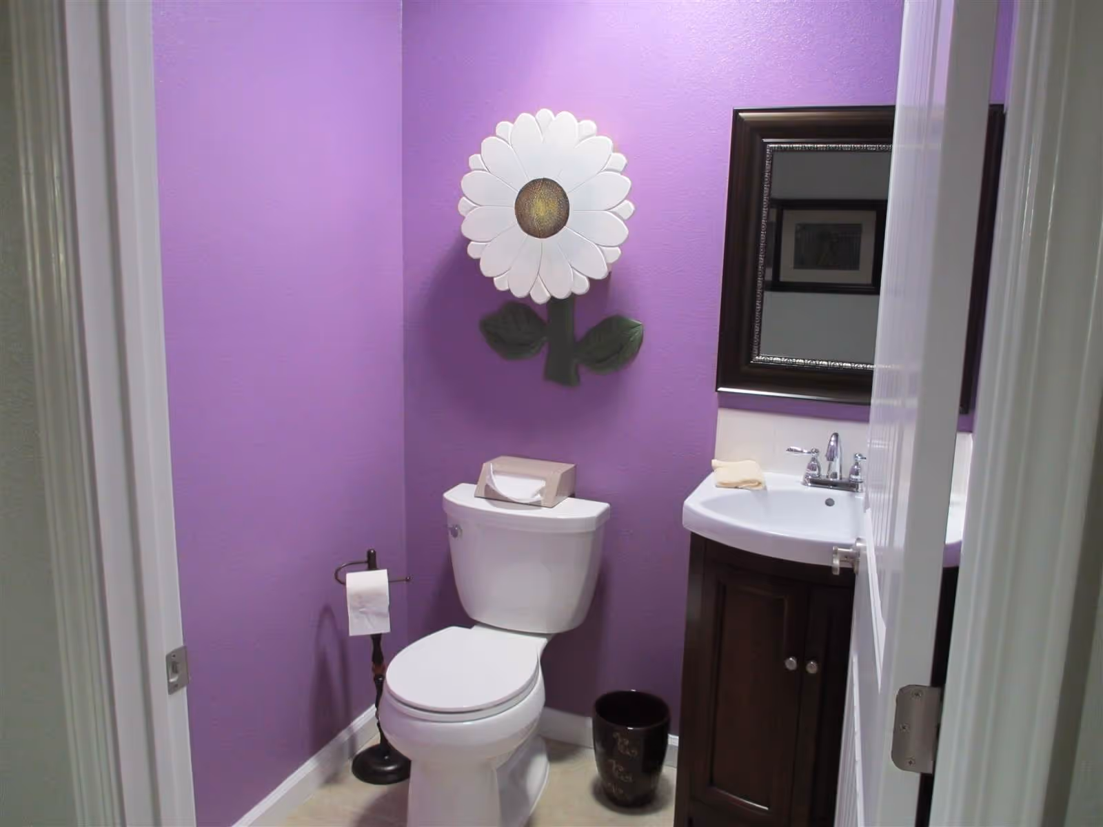 Small bathroom with purple walls featuring a white toilet, dark wood sink vanity, mirror and a large daisy wall decoration.