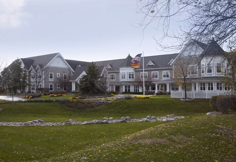 Exterior view of a large senior living facility building with multiple windows, a white picket fence, and a well-maintained lawn with trees and shrubs. An American flag and a Sunrise flag are flying on a flagpole in front of the building under a cloudy sky.