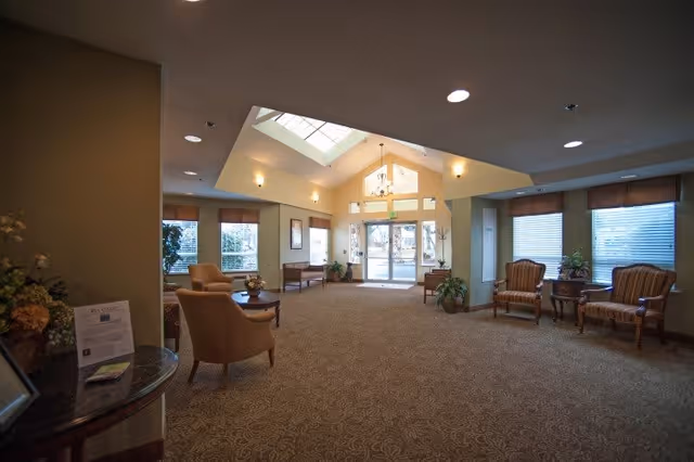 A spacious and well-lit common area with beige carpet and walls, featuring several armchairs and small tables arranged near windows. The ceiling has recessed lighting and a skylight above the entrance door, which has large glass panels allowing natural light to enter. Potted plants and framed pictures decorate the space.