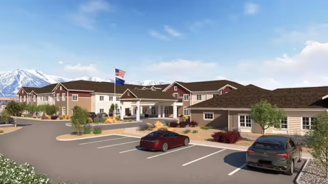 Front exterior of a senior living facility showing the main entrance, a parking lot with two cars, flagpoles, landscaping, and mountains in the background.