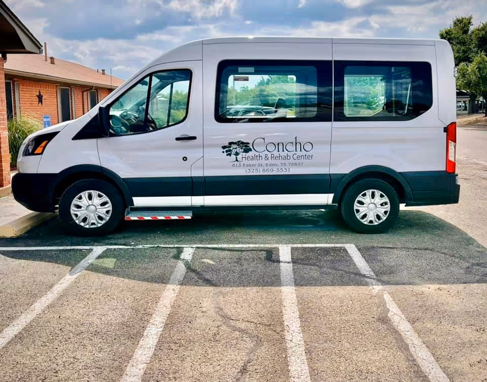 A white passenger van parked in a parking lot next to a brick building. The van has the logo and text for Concho Health & Rehab Center on its side, including the address and phone number.