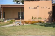 Single-story brick building front reading 'Village Villa 412' with a lawn, walkway, and a bench.