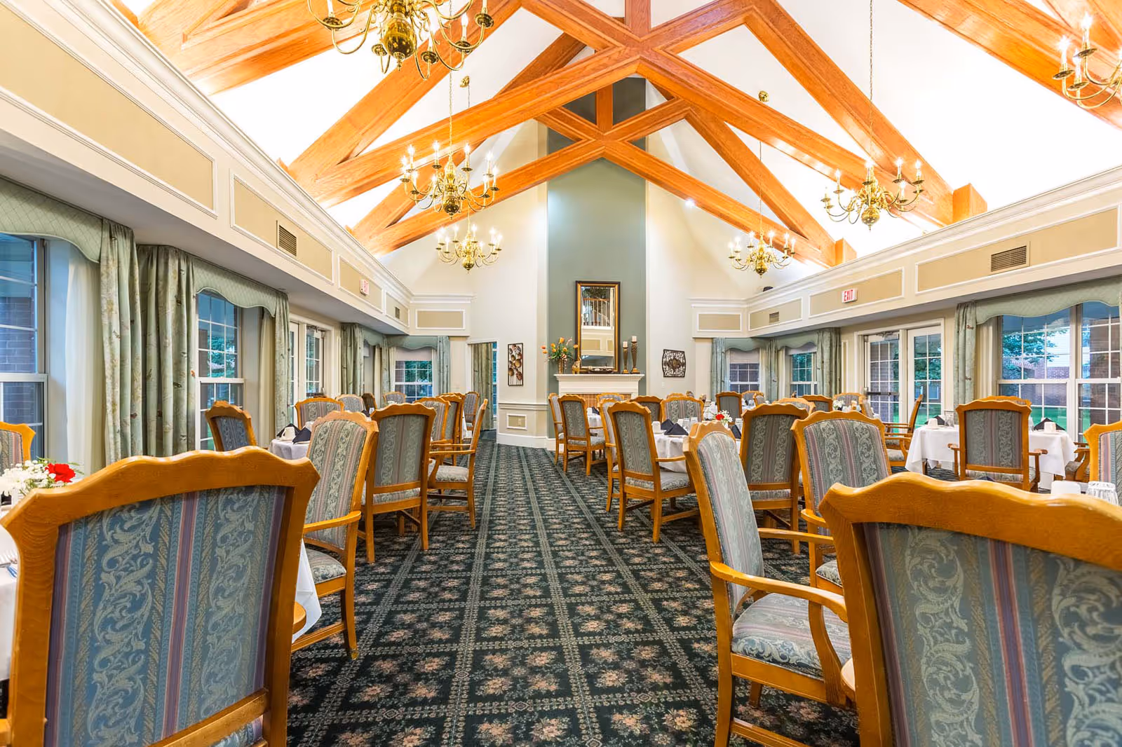 Spacious formal dining room with upholstered wooden chairs and set tables under exposed wooden vaulted beams and chandeliers.