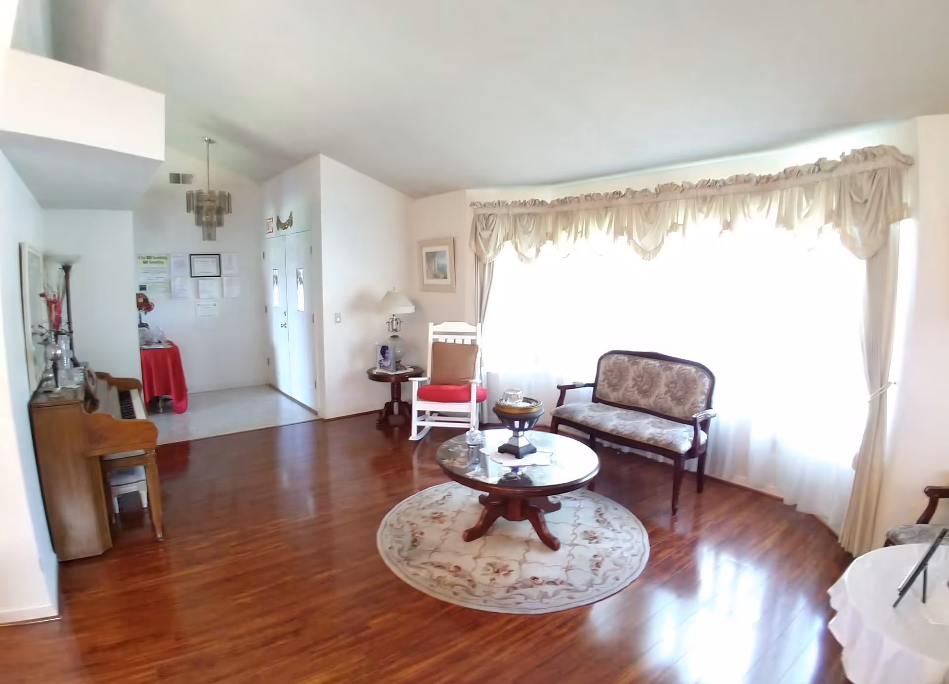 Sunlit living room with hardwood floors, a round coffee table on a rug, an antique settee and chairs, piano and large curtained window.