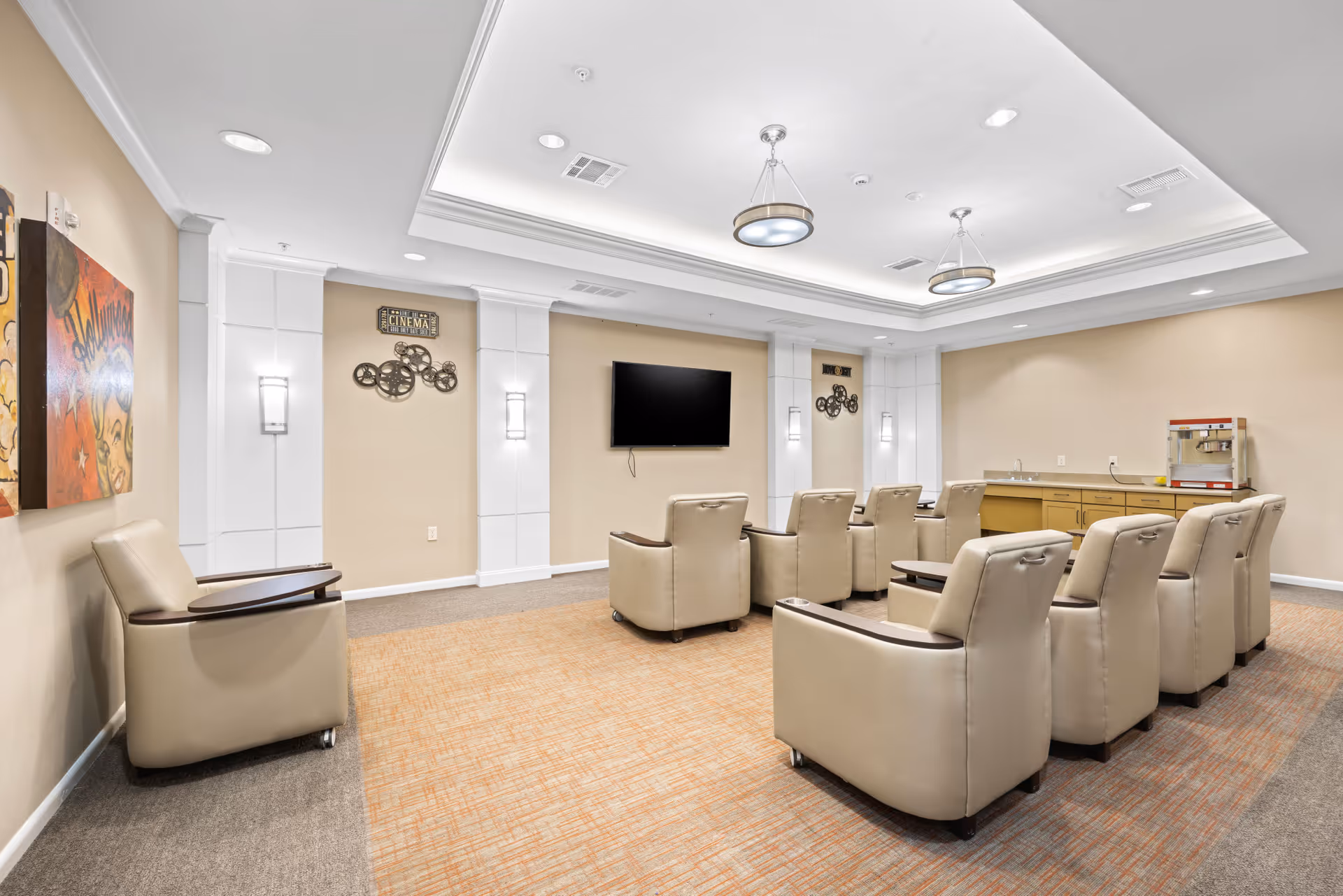A comfortable media room with beige leather recliner chairs arranged in rows facing a wall-mounted flat screen TV. The room has beige walls with white panel accents, modern ceiling lights, and a popcorn machine on a counter in the back corner. There is also colorful artwork on one wall.