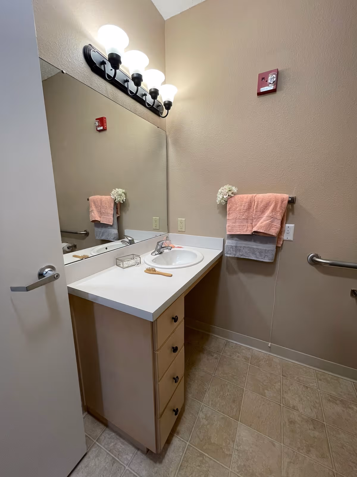 Accessible bathroom vanity with sink, large mirror, row of lights, towel rack holding pink and gray towels, grab bars, and tiled floor.