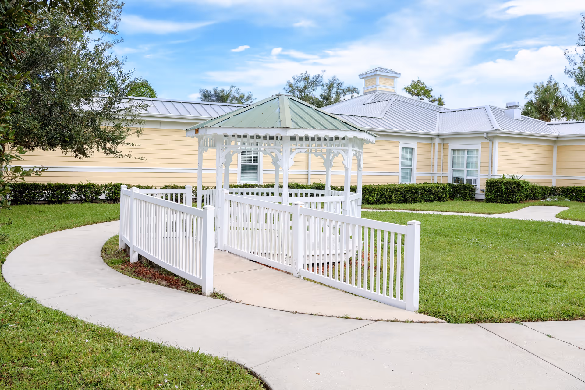 Outdoor view of a senior living facility with a white gazebo featuring a green roof, surrounded by a white fence and a curved concrete pathway. The background shows a yellow building with white trim and a metal roof, along with green grass and trees under a partly cloudy sky.