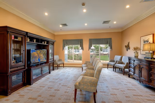 A well-lit living room with beige walls and carpeted floor featuring a row of six upholstered chairs facing a large wooden entertainment center with a flat-screen TV. Two large windows with patterned valances allow natural light to enter. Additional chairs and a wooden sideboard with a lamp and decorative items are placed along the walls.