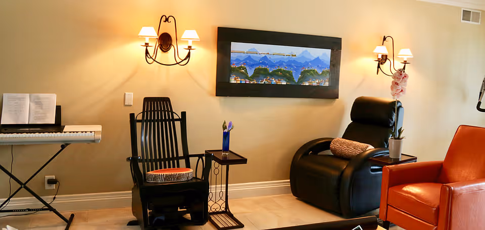 A cozy living room area with a black rocking chair, a black recliner with a pillow, and an orange armchair. There is a small side table with a vase and flowers between the chairs. On the wall above the chairs are two wall-mounted lamps and a framed picture of a mountain landscape. To the left, there is an electronic keyboard with sheet music on it.