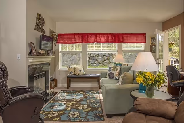 A cozy living room with a green sofa adorned with floral pillows, a brown recliner chair, a patterned area rug with blue and beige flowers, a small round table with a lamp and a vase of yellow flowers, a bench with cushions under three windows with red valances, and a fireplace on the left side.
