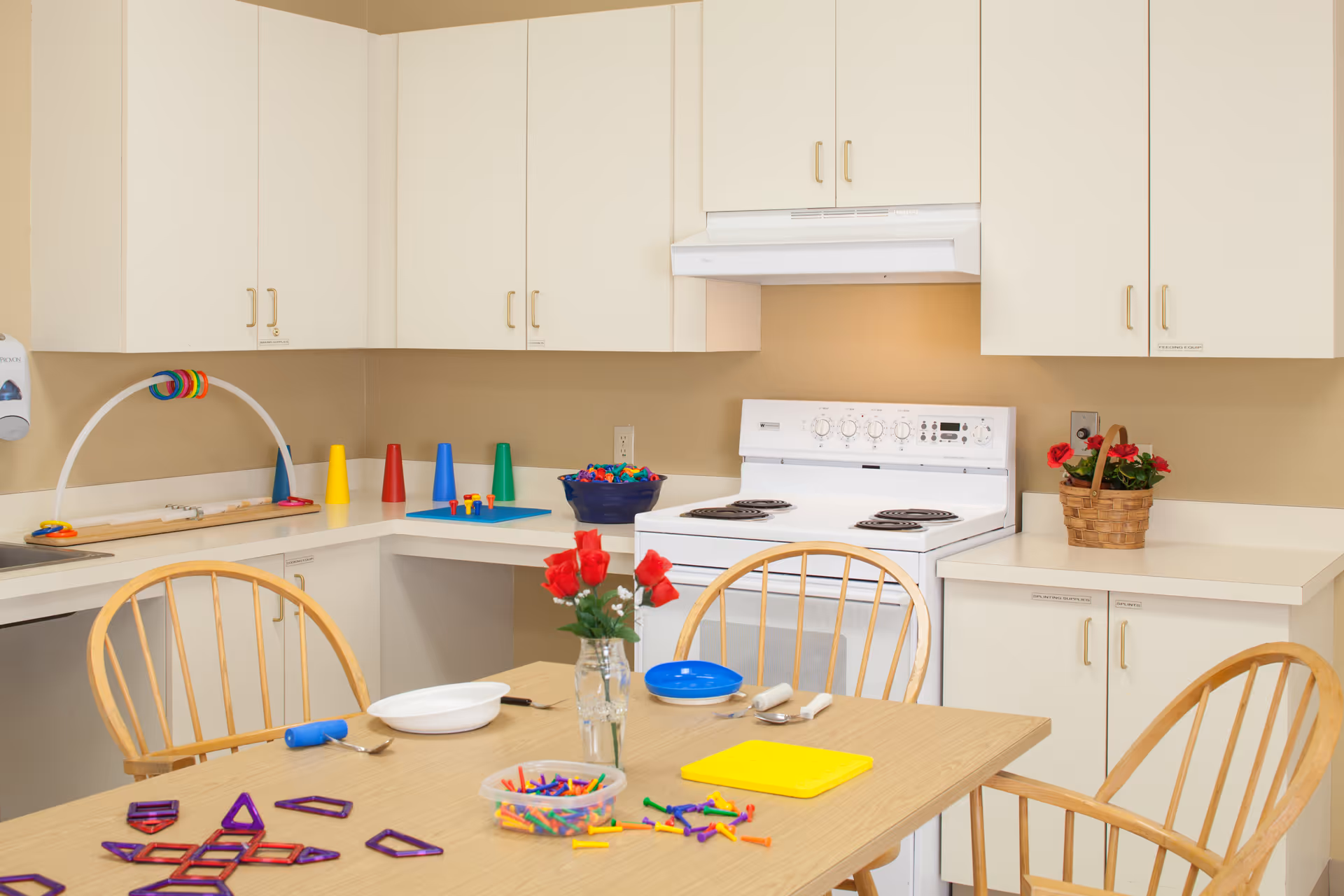 A bright kitchen area with white cabinets and a white electric stove. A wooden table with four chairs is in the foreground, with various colorful toys and craft materials scattered on the table. A small vase with red flowers is also on the table. There is a basket with red flowers on the countertop to the right.