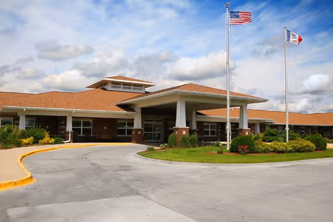 Exterior view of a single-story senior living facility building with a covered entrance, two flagpoles displaying the American flag and another flag, surrounded by landscaped greenery and a circular driveway under a partly cloudy sky.