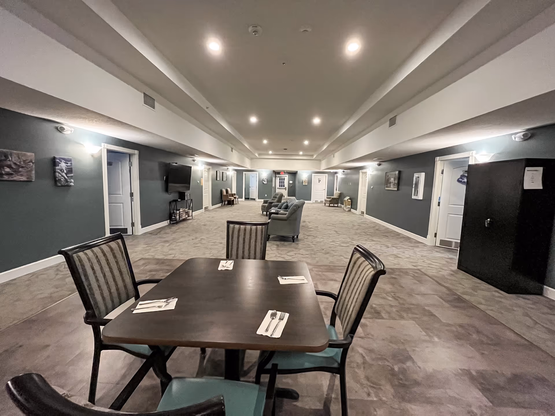 A spacious interior common area in an assisted living facility with a dining table set for four in the foreground and several armchairs arranged along the walls in the background. The walls are painted dark teal with white trim, and the ceiling has recessed lighting. There is a flat-screen TV mounted on the left wall and various framed artworks on the walls.
