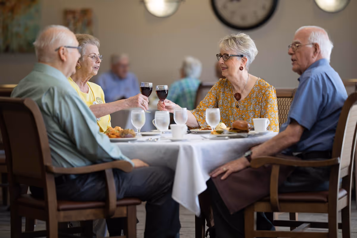 Four elderly people sitting around a dining table in a senior living facility, two women clinking glasses of red wine while the others look on. The table is set with plates of food, glasses of water, and cups. A large clock is visible on the wall in the background.